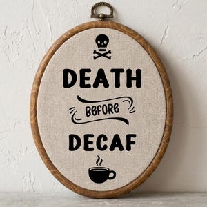 May include: An oval embroidery hoop with a wooden frame containing beige fabric. The text "DEATH BEFORE DECAF" is printed in black, accompanied by a black skull and crossbones icon at the top and a black coffee cup icon at the bottom.