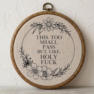 Könnte beinhalten: Ein runder hölzerner Stickrahmen mit beigem Stoff, auf dem der schwarze Text "THIS TOO SHALL PASS BUT, LIKE, HOLY FUCK" gestickt ist, umgeben von einem schwarzen Blumenkranz aus verschiedenen Blüten und Blättern.