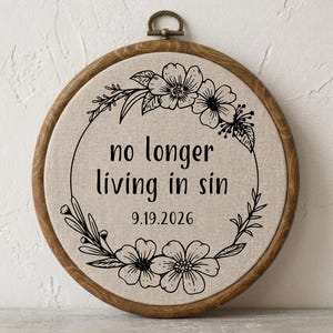 May include: A circular wooden embroidery hoop containing beige fabric with a black floral wreath design. The text "no longer living in sin" is printed in the centre, followed by the date "9.19.2026" in a simple black sans-serif font.