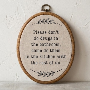 以下が含まれることがあります： ベージュの布地に黒い刺繍で「Please don't do drugs in the bathroom, come do them in the kitchen with the rest of us.」と書かれた楕円形の刺繍枠。フレームはダークウッドの木目調仕上げ。