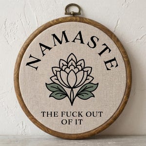 May include: A circular wooden embroidery hoop containing beige fabric with black embroidered text that reads NAMASTE THE FUCK OUT OF IT. Centered between the two lines of text is a black and sage green embroidered lotus flower design.