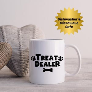 May include: White ceramic mug with the text "TREAT DEALER" in black, featuring paw prints and a bone graphic. A gold starburst sticker indicates the mug is dishwasher and microwave safe.