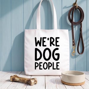 Op de afbeelding: Witte draagtas met de tekst "WE'RE DOG PEOPLE" in het zwart. Een bruine leren riem hangt aan een haak. Een hondenbak en een botvormig kauwspeeltje liggen op een wit houten oppervlak. De achtergrond is een lichtblauwe muur.