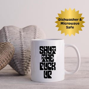 Può includere: Tazza in ceramica bianca con la scritta "SHUT THE FUCK UP" in grassetto nero in stile retrò. La tazza ha un manico curvo ed è posta su uno sfondo neutro. Una grafica a stella dorata indica che la tazza è lavabile in lavastoviglie e adatta al microonde.