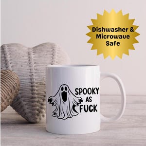Puede incluir: Taza de cerámica blanca con un diseño de fantasma negro y la frase "SPOOKY AS FUCK". La taza tiene un asa curvada y está etiquetada como "Dishwasher & Microwave Safe" en una estrella dorada.