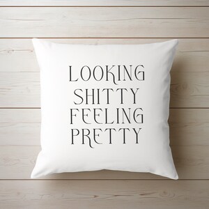 May include: White square pillow with the text "LOOKING SHITTY FEELING PRETTY" in black, centered on the front. The pillow is on a light wood surface.