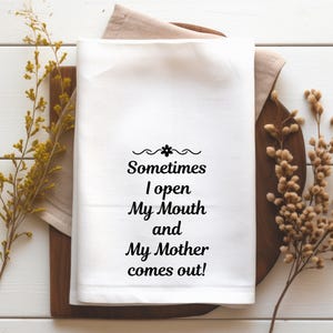 May include: White kitchen towel with the text "Sometimes I open My Mouth and My Mother comes out!" in black script. The towel is on a wooden cutting board with a beige linen napkin underneath. Dried floral accents are on either side.