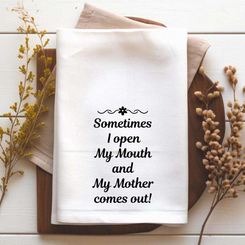May include: White kitchen towel with the text "Sometimes I open My Mouth and My Mother comes out!" in black script. The towel is on a wooden cutting board with a beige linen napkin underneath. Dried floral accents are on either side.