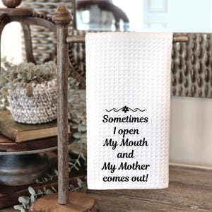 May include: White waffle-weave kitchen towel with the text "Sometimes I open My Mouth and My Mother comes out!" displayed in black script. The towel is hanging on a wooden towel rack.