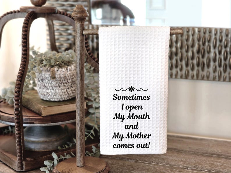 May include: White waffle-weave kitchen towel with the text "Sometimes I open My Mouth and My Mother comes out!" displayed in black script. The towel is hanging on a wooden towel rack.