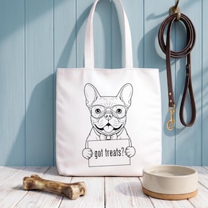 Op de afbeelding: Witte draagtas met een zwart-wit illustratie van een Franse Bulldog met een bril en een vlinderdas, met een bord met de tekst "got treats?". Een bruine leren riem hangt in de buurt. Een hondenbak en bot zijn ook zichtbaar.