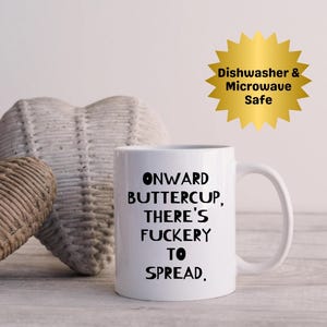 Puede incluir: Taza de cerámica blanca con el texto "ONWARD BUTTERCUP, THERE'S FUCKERTY TO SPREAD". Un gráfico de estrella dorada dice "Dishwasher & Microwave Safe". La taza tiene un asa blanca y está sobre una superficie de madera clara.