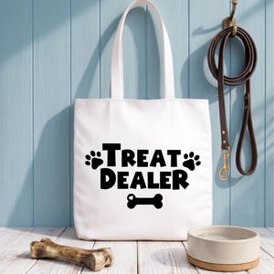 Op de afbeelding: Witte draagtas met de tekst "TREAT DEALER" in zwart, met pootafdrukken en een botafbeelding. Een bruine leren hondenriem hangt aan een haak. Een hondenbak en een bot liggen op een wit houten oppervlak.