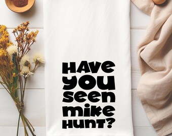 Könnte beinhalten: Weißes Küchentuch mit dem Aufdruck "HAVE YOU SEEN MIKE HUNT?" in fetten schwarzen Buchstaben. Das Handtuch ist gefaltet und liegt auf einer weißen Holzoberfläche, mit dekorativen Elementen im Hintergrund.