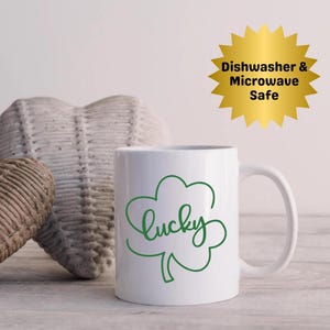 May include: White ceramic mug with a green shamrock design and the word "Lucky" in a cursive font. The mug has a curved handle and the text "Dishwasher & Microwave Safe" on a gold starburst.