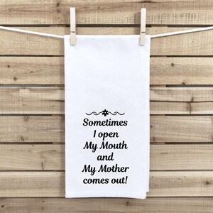 May include: A white kitchen towel with the text "Sometimes I open My Mouth and My Mother comes out!" in black script. The towel is hanging from a clothesline with two wooden clothespins against a wooden background.
