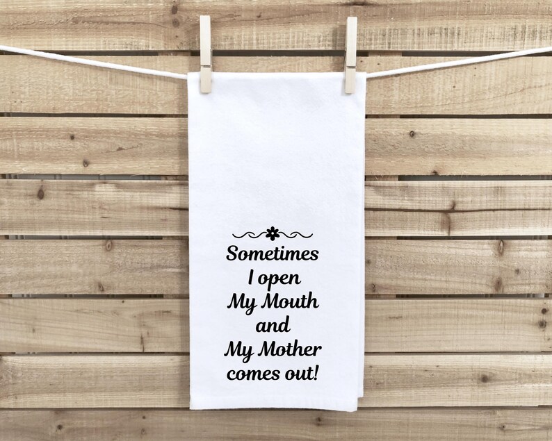 May include: A white kitchen towel with the text "Sometimes I open My Mouth and My Mother comes out!" in black script. The towel is hanging from a clothesline with two wooden clothespins against a wooden background.