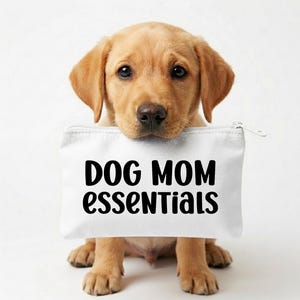 May include: A golden Labrador puppy sits, holding a white zippered pouch. The pouch has the words "DOG MOM essentials" printed in bold black letters. The puppy has a sweet expression.