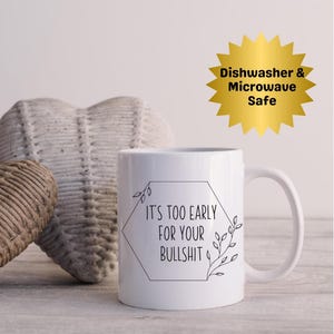 Può includere: Tazza in ceramica bianca con manico curvo. La tazza presenta il testo "It's TOO EARLY FOR YOUR BULLSHIT" all'interno di un esagono nero con accenti di foglie. Una grafica a stella dorata indica che la tazza è lavabile in lavastoviglie e adatta al microonde.