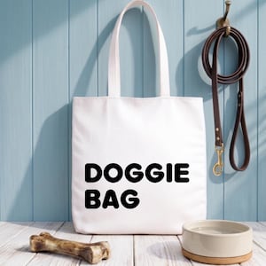 Op de afbeelding: Een witte draagtas met de woorden "DOGGIE BAG" in vette zwarte letters. Een bruine leren hondenriem hangt aan een haak. Een hondenbak en een botvormig kauwspeeltje liggen op een wit houten oppervlak.