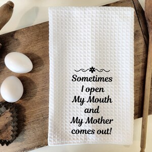 May include: White waffle-weave kitchen towel with the text "Sometimes I open My Mouth and My Mother comes out!" in black. The towel is on a wooden cutting board with eggs, a rolling pin, and a cookie cutter.