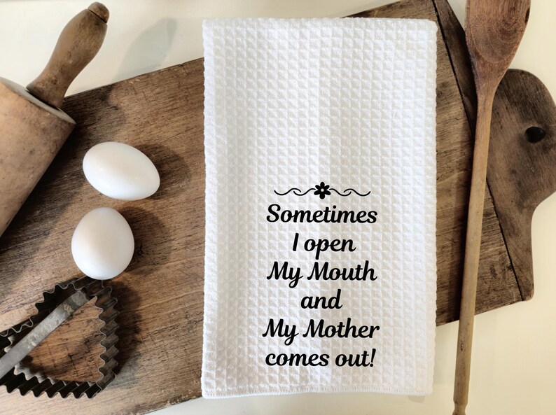May include: White waffle-weave kitchen towel with the text "Sometimes I open My Mouth and My Mother comes out!" in black. The towel is on a wooden cutting board with eggs, a rolling pin, and a cookie cutter.