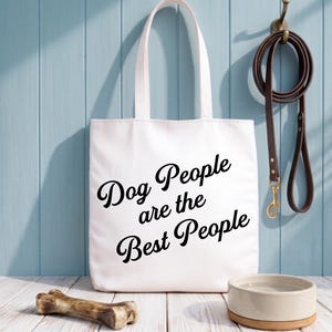 Op de afbeelding: Witte draagtas met de tekst "Dog People are the Best People" in zwart script. Een bruine leren hondenriem hangt aan een haak. Een hondenbot en een beige hondenbak staan op een houten oppervlak.