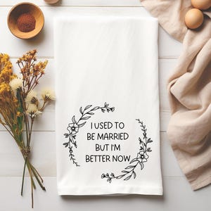 May include: White kitchen towel with the text "I USED TO BE MARRIED BUT I'M BETTER NOW" in black script, surrounded by a floral wreath design. The towel is rectangular and made of a soft fabric.