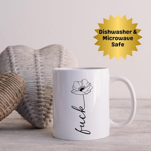 Puede incluir: Taza de cerámica blanca con un diseño floral negro y la palabra "fuck" en cursiva. Un gráfico de estrella dorada indica que la taza es apta para lavavajillas y microondas. La taza tiene un asa curvada.