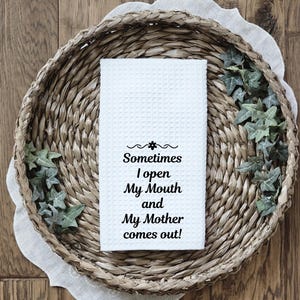 May include: A white waffle-weave kitchen towel with the text "Sometimes I open My Mouth and My Mother comes out!" is displayed in a woven basket with decorative greenery. The towel is centered in the image.