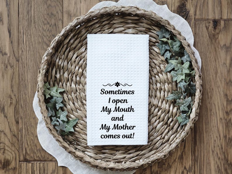 May include: A white waffle-weave kitchen towel with the text "Sometimes I open My Mouth and My Mother comes out!" is displayed in a woven basket with decorative greenery. The towel is centered in the image.