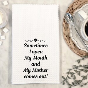 May include: White waffle-weave kitchen towel with the text "Sometimes I open My Mouth and My Mother comes out!" in black script. A cup of coffee, sugar cubes, and a creamer are in the background.