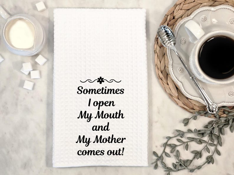 May include: White waffle-weave kitchen towel with the text "Sometimes I open My Mouth and My Mother comes out!" in black script. A cup of coffee, sugar cubes, and a creamer are in the background.