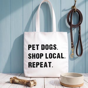 Op de afbeelding: Een witte draagtas met de tekst "PET DOGS. SHOP LOCAL. REPEAT." staat op een wit houten oppervlak. Een bruine leren riem hangt aan een haak, samen met een hondenbak en een bot.