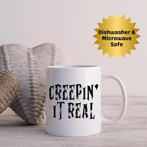 Puede incluir: Taza de cerámica blanca con la frase "CREEPIN' IT REAL" en letras góticas negras. Una pegatina dorada en forma de estrella indica que la taza es apta para lavavajillas y microondas. La taza tiene un asa curvada.