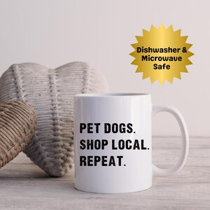 Könnte beinhalten: Weiße Keramik-Tasse mit dem Text "PET DOGS. SHOP LOCAL. REPEAT." in Schwarz. Ein goldener Sternaufkleber mit "Dishwasher & Microwave Safe" ist sichtbar. Zwei herzförmige Webdekorationen befinden sich im Hintergrund.