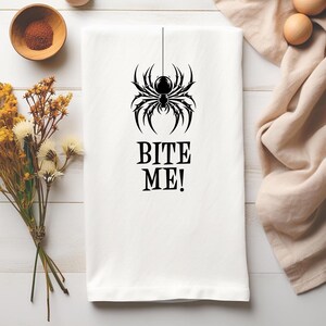 以下が含まれることがあります： 黒いクモのグラフィックと「BITE ME！」のテキストが入った白いキッチンタオル。クモのデザインは、複数の脚と丸い体で詳細に描かれています。タオルはおそらく綿または同様の吸収性素材で作られています。