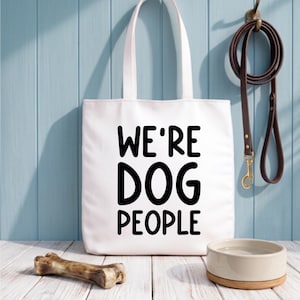Op de afbeelding: Witte draagtas met de tekst "WE'RE DOG PEOPLE" in zwart. Een bruine leren hondenriem hangt aan een haak. Een hondenbak en bot liggen op een houten oppervlak.