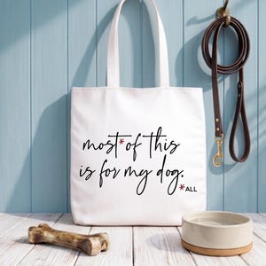 Op de afbeelding: Een witte draagtas met de tekst "most of this is for my dog." Een bruine leren riem hangt aan een haak. Een hondenbot en een hondenbak staan op een houten oppervlak. De achtergrond is een lichtblauwe muur.