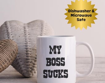 May include: White ceramic mug with the text "MY BOSS SUCKS" in bold black letters. The mug has a curved handle and is set against a neutral background. A gold starburst graphic indicates it is dishwasher and microwave safe.