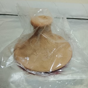 Puede incluir: Un objeto de cerámica hecho a mano, de color marrón claro, posiblemente un pequeño mortero y maja, está encerrado en plástico transparente. El objeto tiene una base redondeada y un mango cilíndrico. La cerámica tiene una superficie texturizada con grietas visibles.