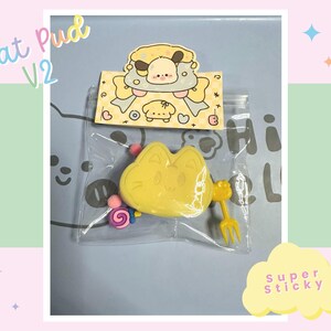May include: A yellow cat-shaped eraser with a fork and a lollipop charm. The eraser is in a clear plastic bag with a sticker that says "Cat Pud V2" and "Super Sticky".