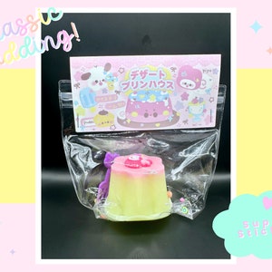 May include: A green and pink pudding-shaped squishy toy with a purple bow and sprinkles. The toy is in a clear plastic bag with a pink and white card that says "Dessert Pudding" in Japanese.
