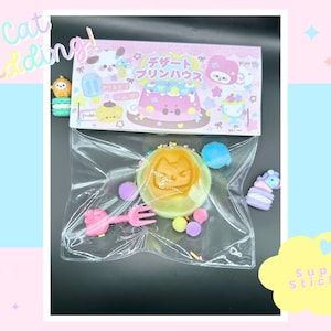 May include: A clear plastic bag containing a yellow cat-shaped pudding, pink and blue sprinkles, a pink fork, a blue and white unicorn-shaped charm, and a pink and white cat-shaped charm. The bag is labelled "Sweet Pudding" in Japanese.