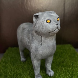 May include: A gray wooden sculpture of a Scottish Fold cat with yellow eyes. The cat is standing on a green surface.