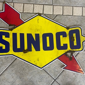May include: A vintage Sunoco petrol station sign with a yellow diamond shape and a red arrow. The sign has the word "SUNOCO" in blue letters.