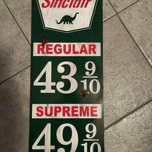 May include: A vintage Sinclair petrol station sign with a green background and white lettering. The sign features a dinosaur logo and lists the prices for regular and supreme petrol as 43 9/10 and 49 9/10.