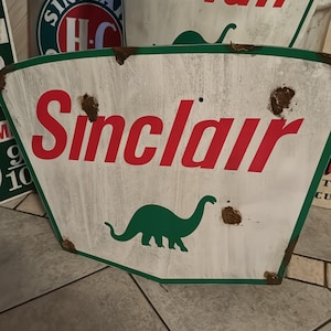 May include: Vintage Sinclair gas station sign with a green dinosaur and the text "Sinclair" in red letters. The sign is white with green and red accents and has some rust spots.