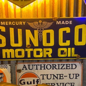 May include: A vintage Sunoco Motor Oil sign with yellow lettering on a dark blue background. The sign features the word "SUNOCO" prominently, with "MOTOR OIL" below. The sign has a weathered appearance, suggesting age and use. The sign is mounted on a corrugated metal surface.