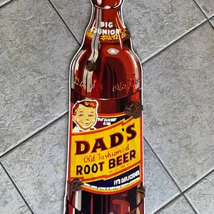 May include: A retro-style sign in the shape of a root beer bottle. The sign displays the "Dad's Old Fashioned Root Beer" logo in yellow and red, with "Big Junior" at the top. The bottle is brown with a gold cap.